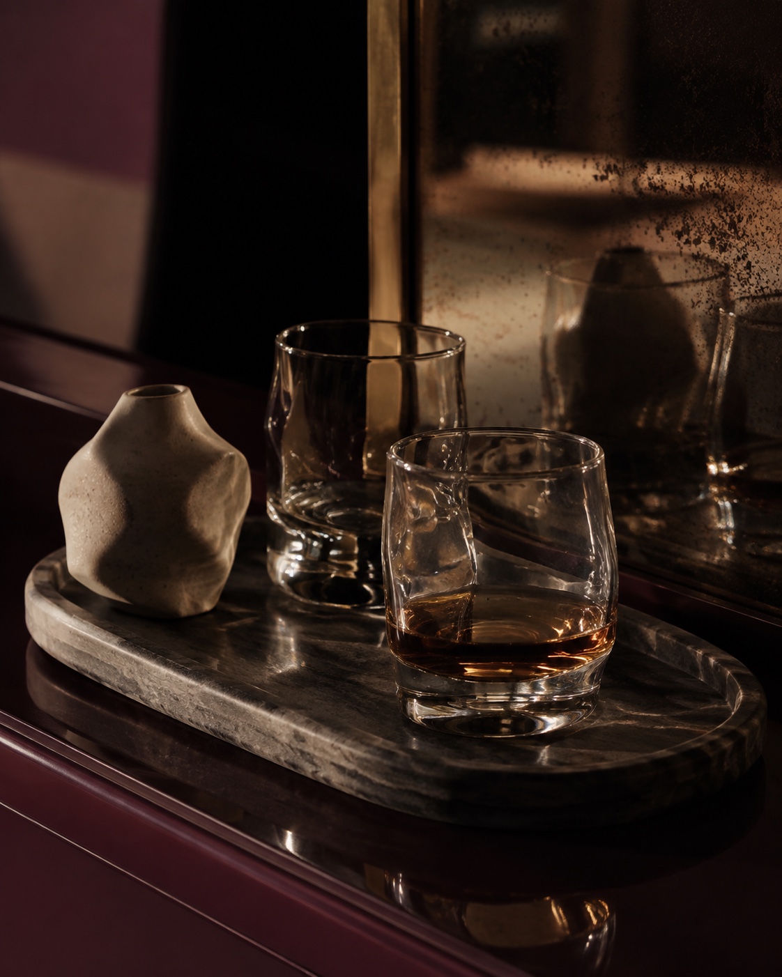 Sculptural glasses and a small ceramic vessel on a veined stone tray, reflected in an aged mirror above an oxblood cabinet